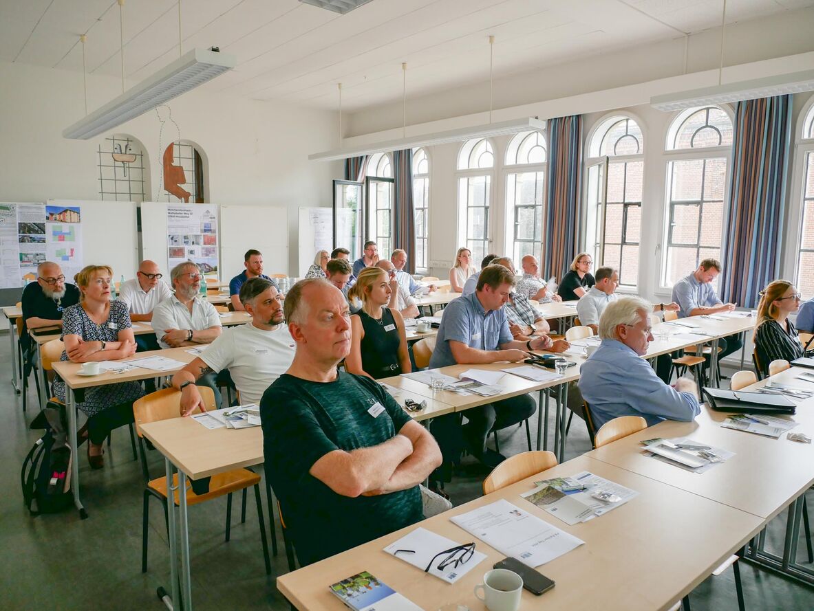 In einem Hörsaal sitzen die Praxispartner an Tischen und blicken Richtung Leinwand