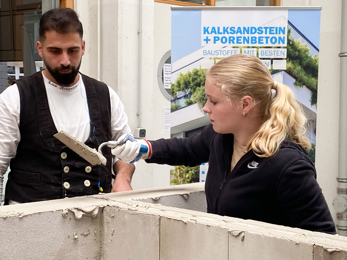 Eine Studentin spachtelt unter Aufsicht eines Experten Mörtel auf das Mauerwerk