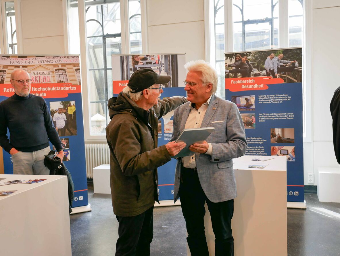 Links steht Professor Stura und überreicht dem rechts stehenden Hochschulpräsidenten Prof. Dr.-Ing. Ingo Hadrych ein Buch als Geschenk.