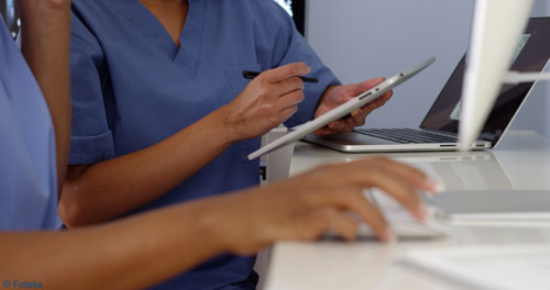 Hände von zwei Pflegefachkräften in blauer Arbeitskleidung, eine Person hält ein Tablet, die andere hält eine Computermaus